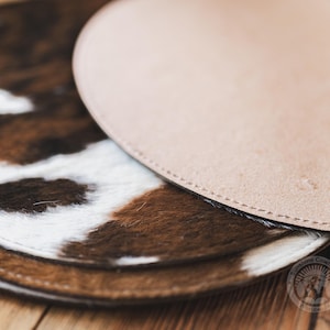 May include: Two circular coasters featuring a brown and white cowhide pattern on the top surface with stitched edges. The underside of the top coaster is made of smooth, light tan leather. A circular logo is visible in the bottom right corner.