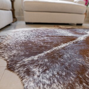 May include: A brown and white cowhide rug with a natural pattern. The rug is oval-shaped and has a soft, textured appearance. The rug is placed on a light-coloured tile floor, with furniture in the background.