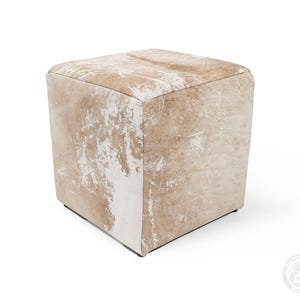 Puede incluir: Un taburete de forma cuadrada tapizado en piel de vaca de color tostado claro y blanco. La superficie presenta un patr&oacute;n natural y moteado con diversos tonos de beige y crema, creando una apariencia texturizada en todos los lados visibles.