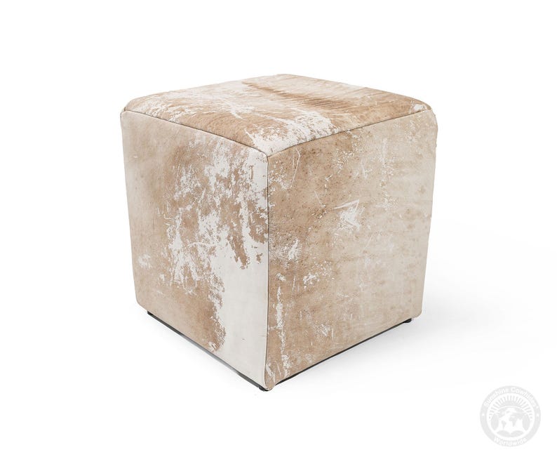 Puede incluir: Un taburete de forma cuadrada tapizado en piel de vaca de color tostado claro y blanco. La superficie presenta un patr&oacute;n natural y moteado con diversos tonos de beige y crema, creando una apariencia texturizada en todos los lados visibles.