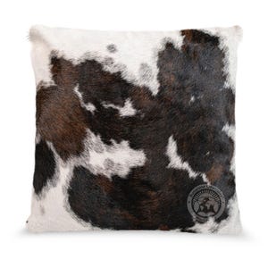 May include: A square throw pillow featuring a natural cowhide pattern with irregular patches of dark brown, black, and white fur. The bottom right corner displays a circular logo with the text Sunshine Cowhides Worldwide.