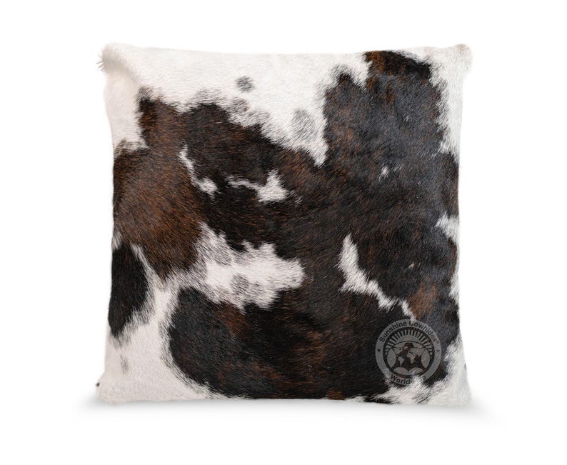 May include: A square throw pillow featuring a natural cowhide pattern with irregular patches of dark brown, black, and white fur. The bottom right corner displays a circular logo with the text Sunshine Cowhides Worldwide.