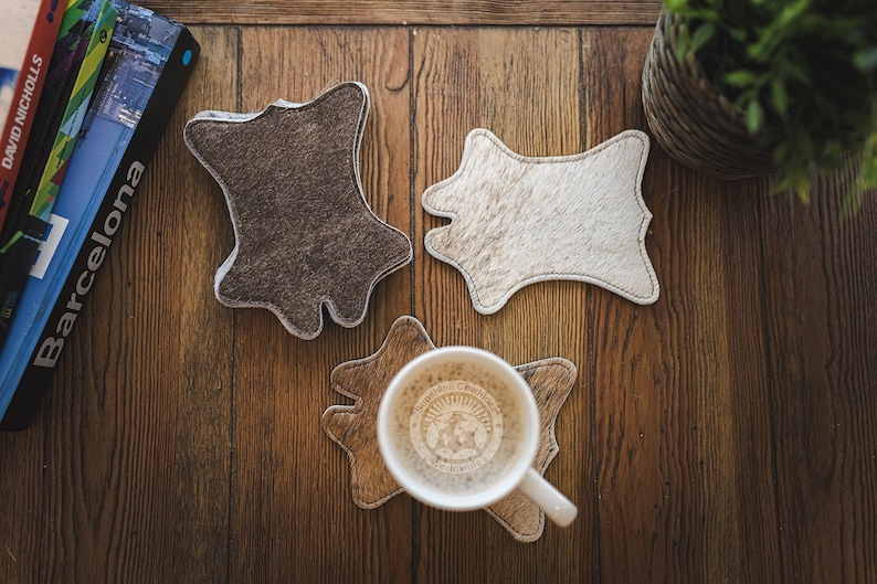 May include: Three cowhide coasters in brown, white, and brown colors. The coasters are in the shape of a cowhide and have a stitched edge. A white coffee mug with a logo is on top of one of the coasters.