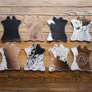 May include: Eight cowhide coasters in shades of black, brown, and white. Each coaster has a distinctive shape with a top cutout. They are displayed on a wooden surface, highlighting their natural textures and colours. The coasters are designed to protect surfaces.