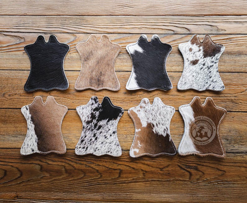 May include: Eight cowhide coasters in shades of black, brown, and white. Each coaster has a distinctive shape with a top cutout. They are displayed on a wooden surface, highlighting their natural textures and colours. The coasters are designed to protect surfaces.