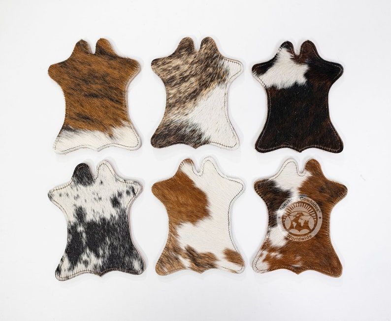 May include: Six cowhide coasters in shades of brown, black, and white. Each coaster has a distinctive shape with a stitched edge. One coaster displays the logo "American Cowhides Worldwide."
