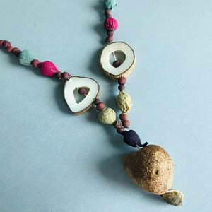 May include: A necklace made of natural materials, including wood, seeds, and dried fruit. The necklace features a variety of colors, including brown, green, red, and white. The necklace is strung on a brown cord and has a large, brown seed at the bottom.