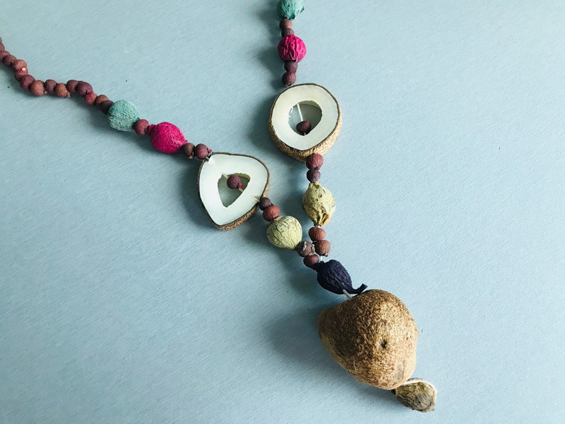 May include: A necklace made of natural materials, including wood, seeds, and dried fruit. The necklace features a variety of colors, including brown, green, red, and white. The necklace is strung on a brown cord and has a large, brown seed at the bottom.