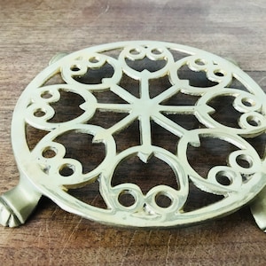 Puede incluir: Un posa-ollas redondo de latón con un diseño decorativo de círculos y hojas superpuestos. El posa-ollas tiene tres patas.