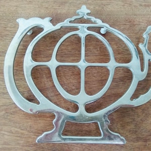 Puede incluir: Salvamanteles de plata con forma de tetera con un diseño decorativo calado. El salvamanteles tiene una base redonda y un asa.