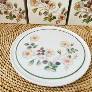 Puede incluir: Un soporte para tartas de cerámica blanca con un borde verde y un diseño floral con flores rosas, amarillas y verdes. El soporte está sobre una superficie de mimbre tejido.