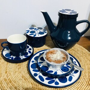 K&ouml;nnte beinhalten: Ein blaues und wei&szlig;es Teeservice mit floralem Muster, bestehend aus Teekanne, Zuckerdose, Tasse und Untertasse sowie einem kleinen Teller mit einem Geb&auml;ck.