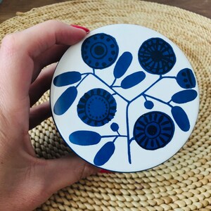 K&ouml;nnte beinhalten: Eine wei&szlig;e Keramik-Schmuckdose mit einem blauen Blumenmuster. Das Design zeigt stilisierte Blumen mit blauen und schwarzen Zentren und blauen Bl&auml;ttern.