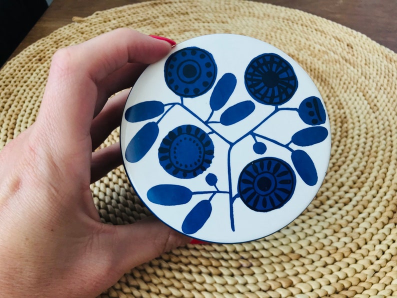K&ouml;nnte beinhalten: Eine wei&szlig;e Keramik-Schmuckdose mit einem blauen Blumenmuster. Das Design zeigt stilisierte Blumen mit blauen und schwarzen Zentren und blauen Bl&auml;ttern.