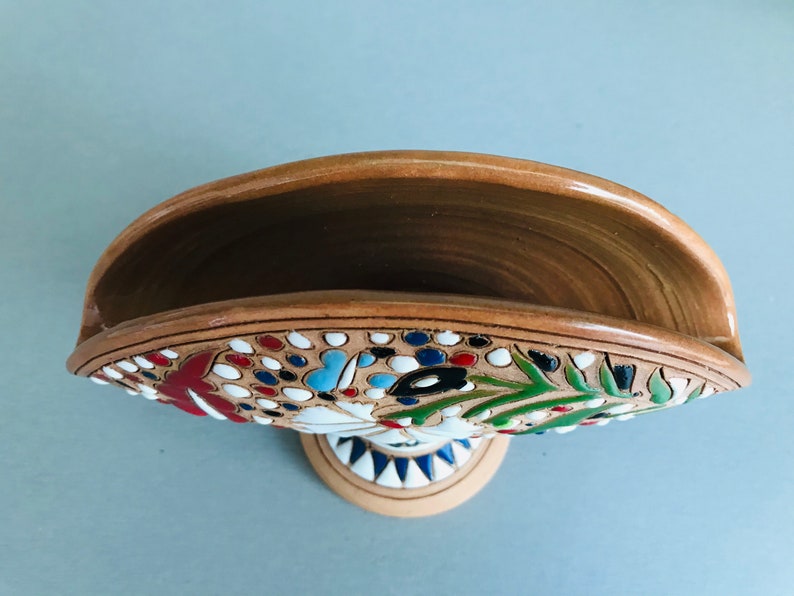 Pode incluir: Um porta-guardanapos de cer&acirc;mica com um design floral em tons de vermelho, azul, verde e branco. O porta-guardanapos tem uma base castanha e uma borda branca.