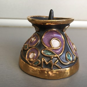 May include: A black ceramic candle holder with a gold rim and purple and green floral designs. The holder has a small black knob on top.