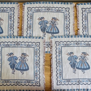 Può includere: Set di sei tovagliette all'americana in tessuto ricamate in blu e bianco con una scena di danza popolare tradizionale. Ogni tovaglietta ha uno sfondo bianco con un bordo blu e un quadrato centrale con due figure in costume tradizionale che ballano.