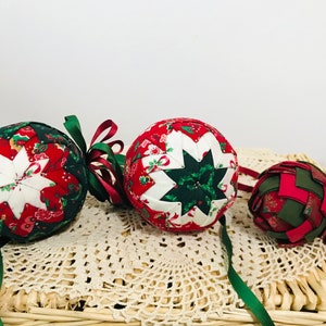 Puede incluir: Tres adornos navideños hechos a mano de tela en rojo, verde y blanco. Los adornos tienen forma de bolas y tienen un diseño de estrella en el centro. Están decorados con cintas.