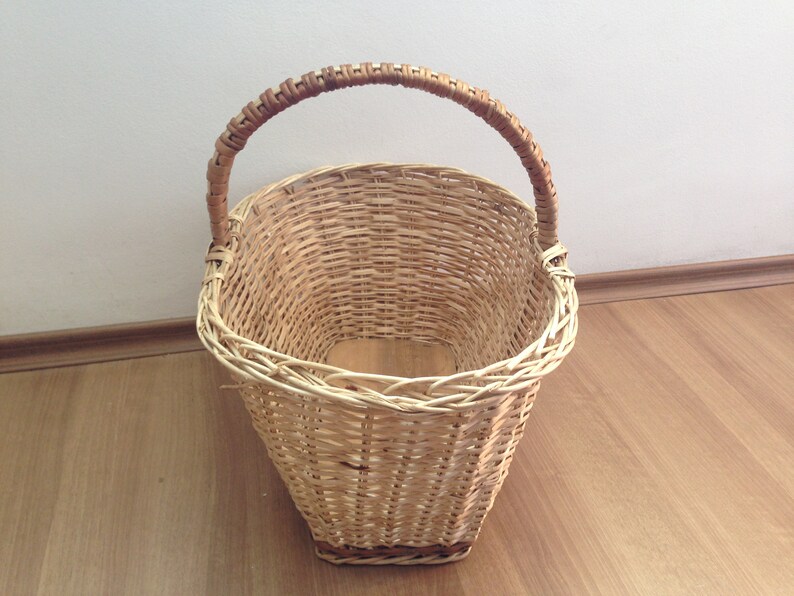 May include: A woven wicker basket with a brown handle. The basket is oval shaped and has a wide opening.