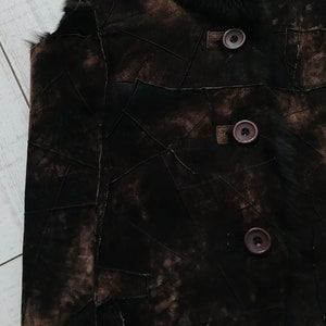 May include: A brown suede vest with a fur collar and three brown buttons. The vest is made of multiple pieces of suede sewn together in a patchwork design.