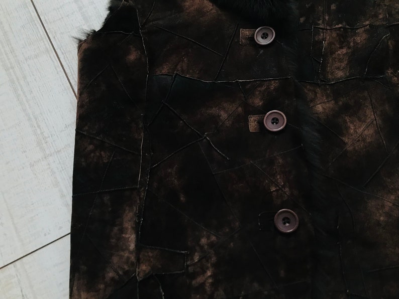 May include: A brown suede vest with a fur collar and three brown buttons. The vest is made of multiple pieces of suede sewn together in a patchwork design.