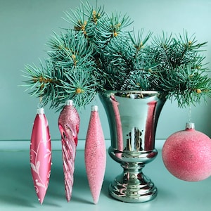 May include: A silver vase with a blue spruce tree inside. Four pink glass ornaments hang from the branches. The ornaments are different shapes and have a sparkly finish.