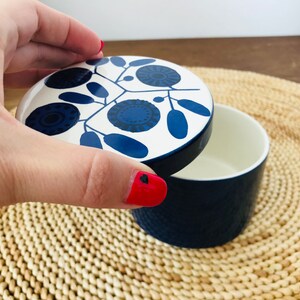 K&ouml;nnte beinhalten: Eine wei&szlig;e Keramikdose mit einem blauen und wei&szlig;en floralen Deckel. Die Dose ist geschlossen und der Deckel wird von einer Hand gehalten.