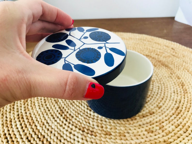 K&ouml;nnte beinhalten: Eine wei&szlig;e Keramikdose mit einem blauen und wei&szlig;en floralen Deckel. Die Dose ist geschlossen und der Deckel wird von einer Hand gehalten.