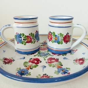 May include: Two white ceramic mugs with blue and red floral designs, sitting on a white ceramic plate with a blue rim and floral designs.