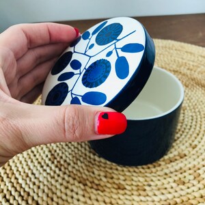 K&ouml;nnte beinhalten: Ein wei&szlig;er Keramikbeh&auml;lter mit einem blauen Deckel. Der Deckel hat ein Blumenmuster mit blauen und schwarzen Akzenten. Der Beh&auml;lter steht auf einer geflochtenen Matte.