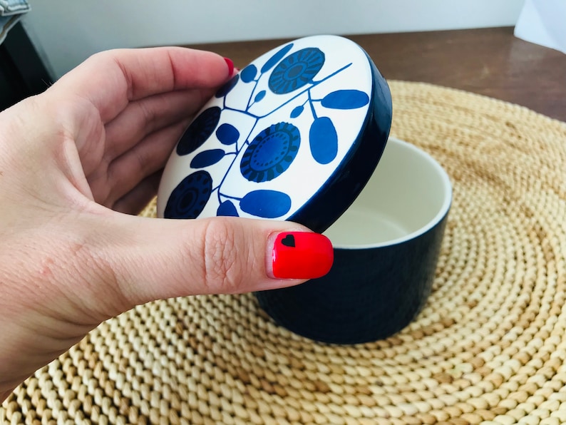 K&ouml;nnte beinhalten: Ein wei&szlig;er Keramikbeh&auml;lter mit einem blauen Deckel. Der Deckel hat ein Blumenmuster mit blauen und schwarzen Akzenten. Der Beh&auml;lter steht auf einer geflochtenen Matte.