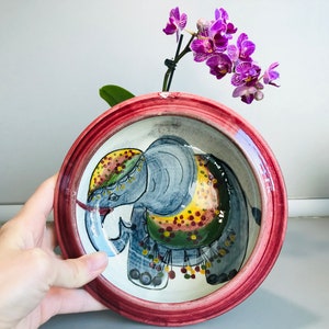May include: A red and white ceramic bowl with a hand-painted design of a gray elephant with yellow, green, and red accents. The bowl is perfect for serving snacks or holding small items.