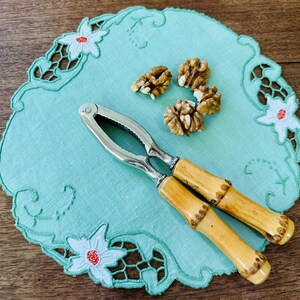 Puede incluir: Un cascanueces de plata con mangos de bambú sobre un mantelito azul claro con flores bordadas blancas. Hay varias nueces en el mantelito.