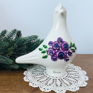 Puede incluir: Figurita de paloma de cerámica blanca con diseño floral morado y verde. La paloma está de pie sobre un encaje blanco. La base de la paloma está inscrita con el texto "Design by Rose Living".