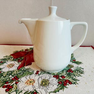 Puede incluir: Jarrita o jarra de cerámica blanca con tapa. La jarrita está sobre un mantel bordado blanco y rojo con un diseño floral.