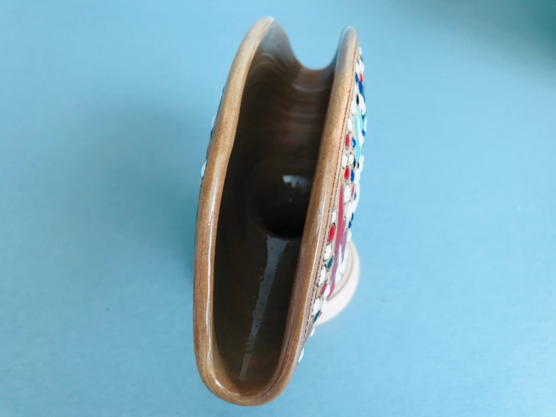 Pode incluir: Um porta-guardanapos de cer&acirc;mica com um esmalte marrom e um design de mosaico com detalhes em vermelho, branco e azul. O porta-guardanapos tem a forma de uma lua crescente e uma abertura larga para os guardanapos.
