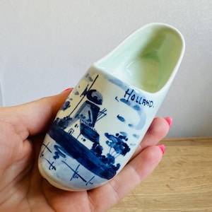May include: A ceramic Dutch clog, painted with a blue and white windmill scene and the word "Holland." The clog is a light blue colour on the inside and is held in a hand.