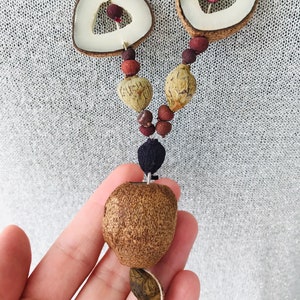 May include: A brown and white seed necklace with a large brown seed pendant. The necklace is made of various natural materials and has a rustic, bohemian style.