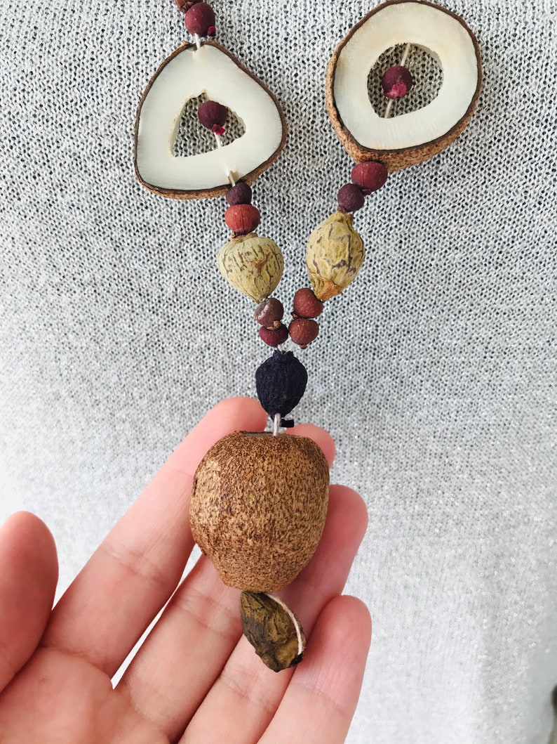 May include: A brown and white seed necklace with a large brown seed pendant. The necklace is made of various natural materials and has a rustic, bohemian style.