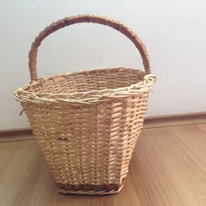 May include: A woven wicker basket with a brown handle. The basket is oval shaped and has a light brown color.
