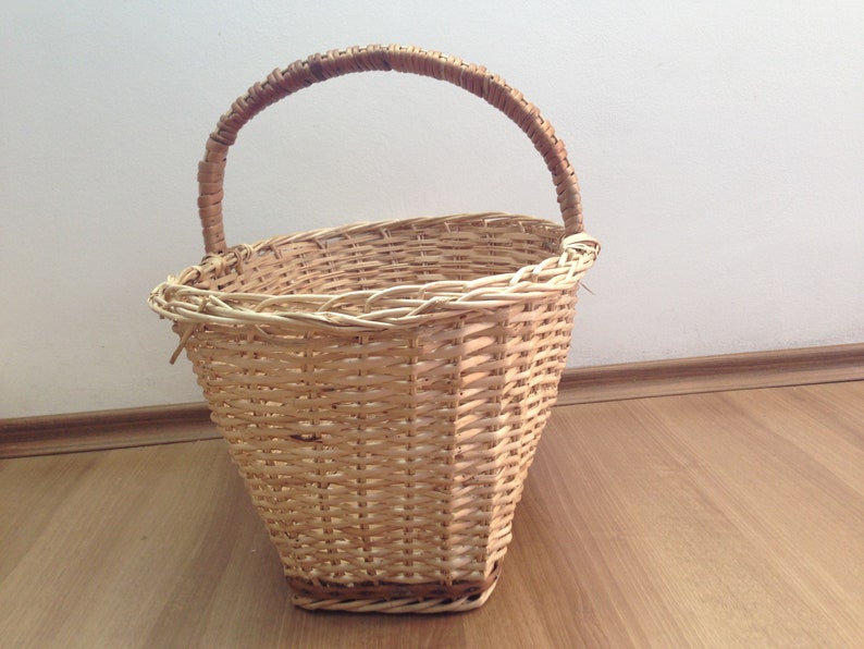May include: A woven wicker basket with a brown handle. The basket is oval shaped and has a light brown color.