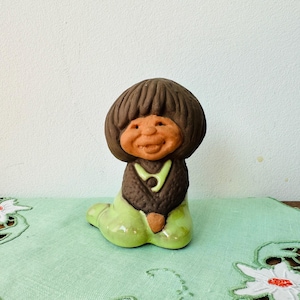 May include: A ceramic figurine of a smiling person with brown hair and a green dress. The figurine is sitting on a green and white lace tablecloth.