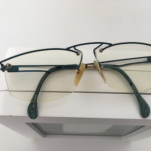 May include: A pair of rimless glasses with a dark green metal frame and gold accents. The lenses are clear and the arms are dark green with a wood grain pattern.