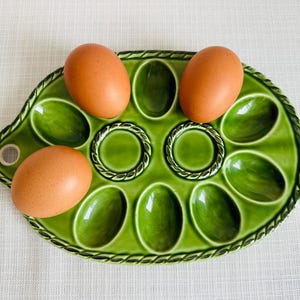 May include: A green ceramic egg holder with a decorative rope-like border, holding four brown eggs. The egg tray has oval and round compartments. The eggs are a light brown colour.