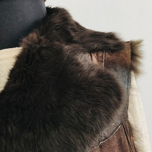 May include: A brown suede vest with a large, fluffy fur collar. The vest is sleeveless and has a zipper closure.