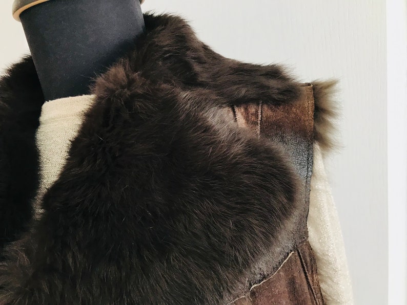 May include: A brown suede vest with a large, fluffy fur collar. The vest is sleeveless and has a zipper closure.