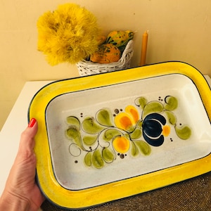 May include: A white ceramic rectangular baking dish with a yellow rim and a black floral design featuring green leaves, yellow and orange flowers, and a blue flower.