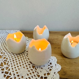 Może przedstawiać: Cztery białe ceramiczne świeczniki w kształcie skorupki jajka ze świecami. Świece emitują ciepłą, żółtą poświatę. Świeczniki są ułożone na drewnianej powierzchni z białą koronkową serwetką.