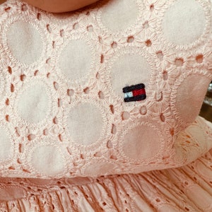 May include: A pale pink dress with a delicate eyelet pattern and a small embroidered logo on the front.