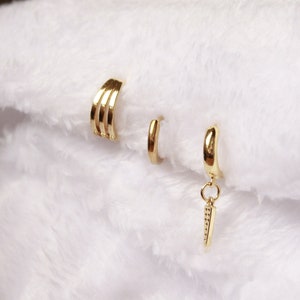 Op de afbeelding: Drie gouden oorringen, waarvan één met een hangende hanger. De hanger is een kleine, gouden, ruitvormige hanger.